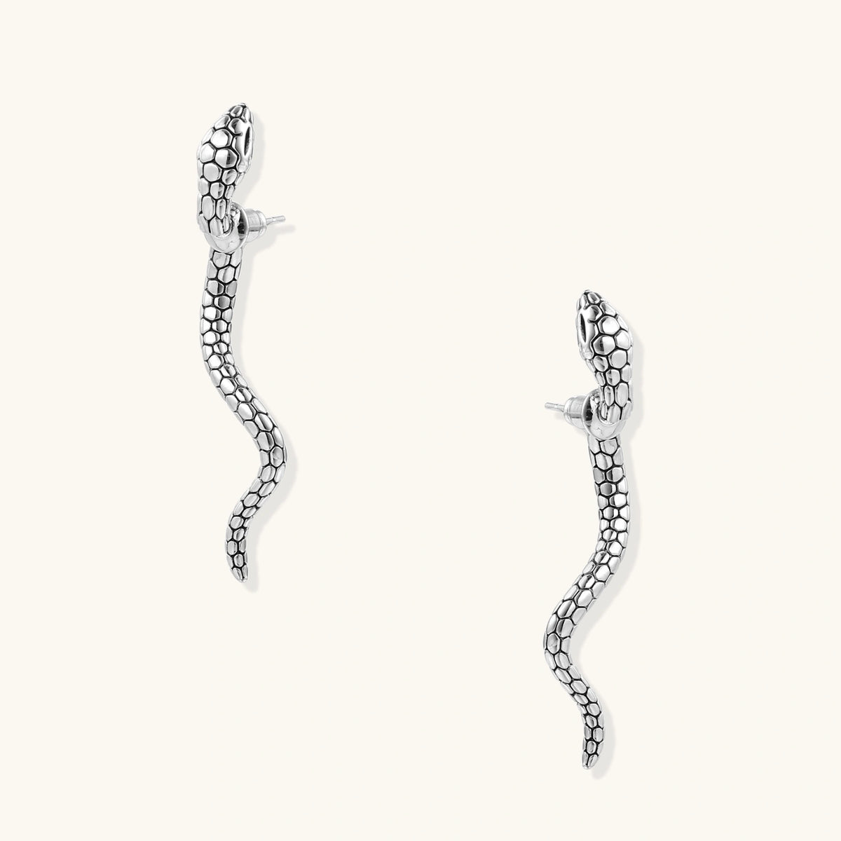 Silver-toned serpent-shaped dangle earrings with detailed scale texture curving in S-shape on beige background