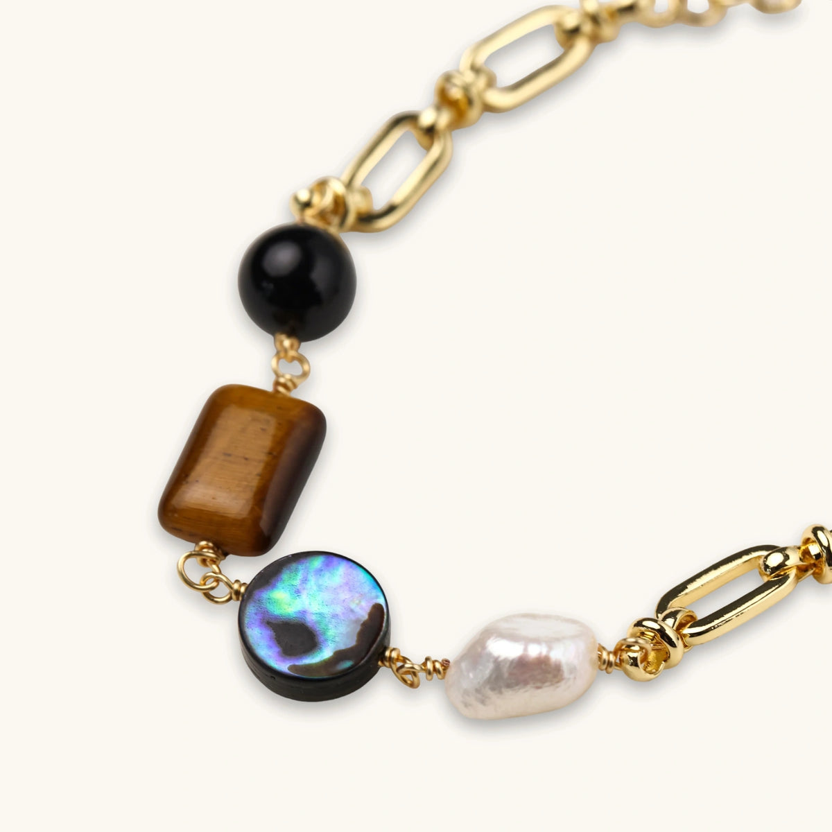 Gold chain bracelet with tiger's eye, obsidian, pearl, and iridescent abalone stones on cream background.