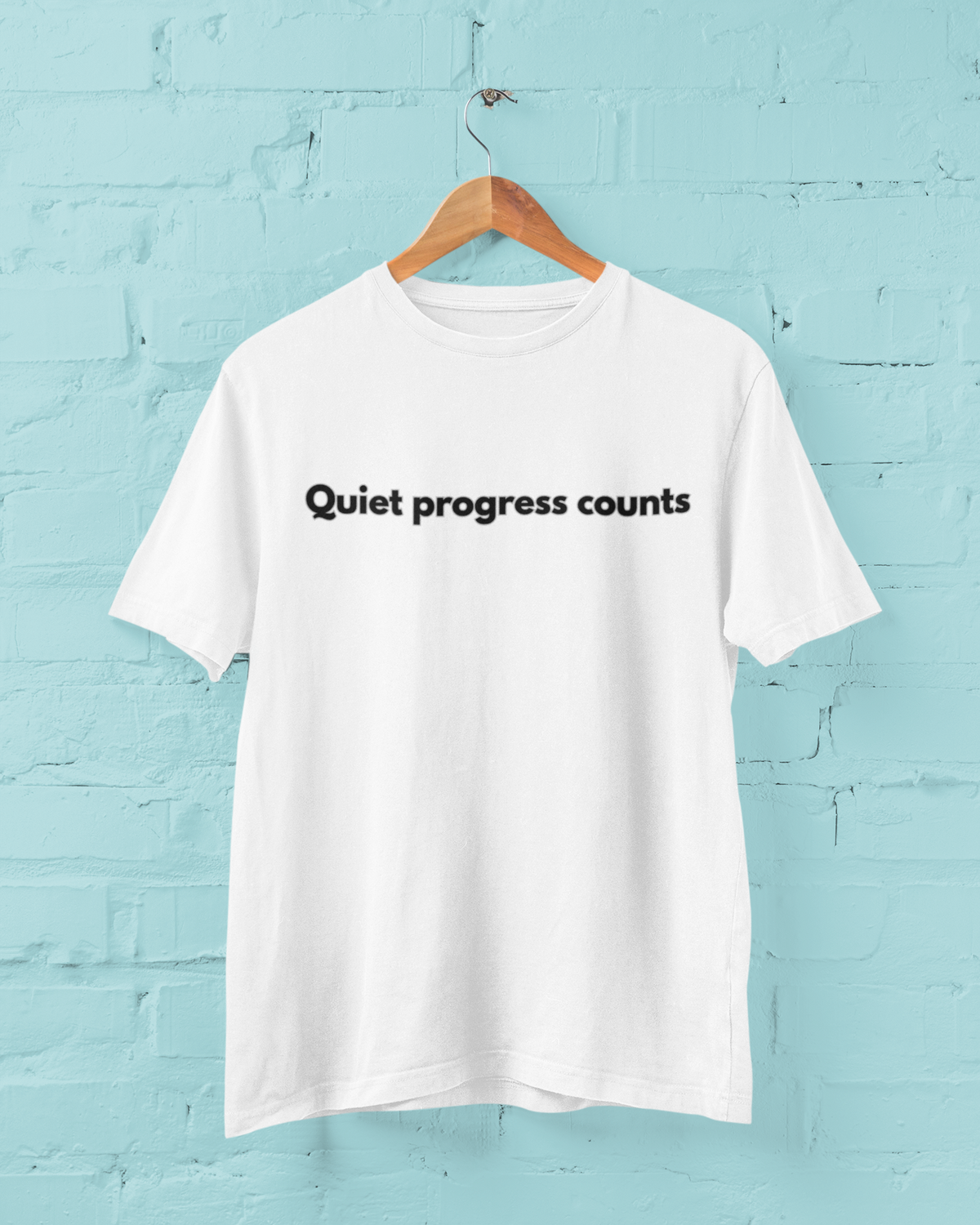 White T-shirt on a wooden hanger against a pale blue wall, with Quiet progress counts printed in black across the chest.